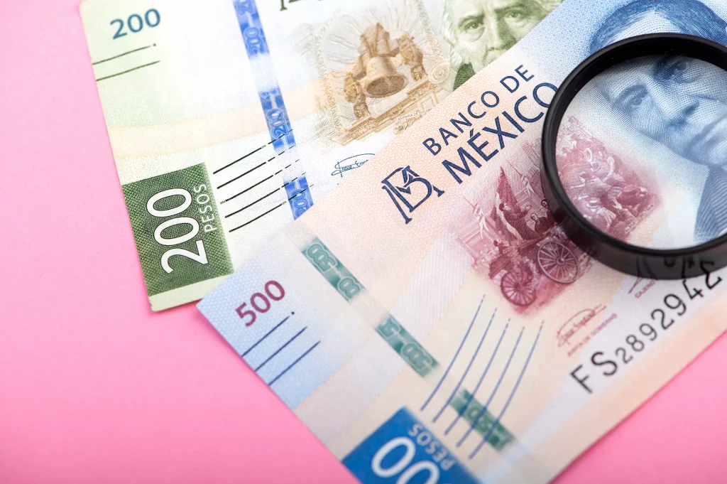 Pesos mexicanos de diferentes denominaciones con lupa sobre fondo rosa, símbolo de análisis financiero e inversión inteligente en México.