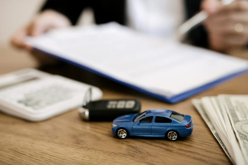 Persona revisa cuentas y documentación junto a una calculadora, dinero y un modelo de auto. La imagen representa la evaluación entre seguir ahorrando para el enganche o recurrir a un préstamo para avanzar con la compra. 
